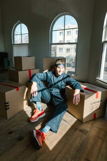 A young man in casual clothing, including a headband, navy blue sweatshirt, and orange running shoes, sitting on a wooden floor surrounded by several large cardboard moving boxes with red and black pa