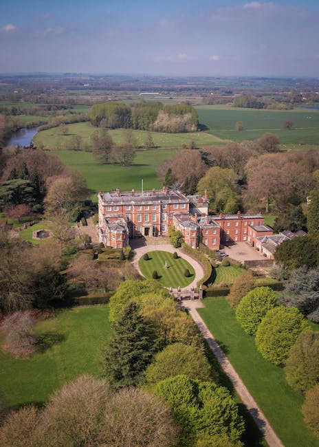 An aerial view of a large, historic brick mansion with multiple wings and decorative architectural features, situated within well-maintained gardens and surrounded by lush green lawns, trees, and a winding pathway leading up to the entrance. Beyond the property, the landscape extends into rolling fields, woodlands, and a river visible in the distance, indicating a rural setting near Forty Hall landmarks. The area appears tranquil with partly cloudy skies overhead, and no moving activity or furniture is visible in the image, focusing solely on the scenic environment associated with house removals and property relocation near Enfield. Enfield Removals may utilize such estates for context in their house removal services, emphasizing meticulous packing and transportation processes within large, stately homes.