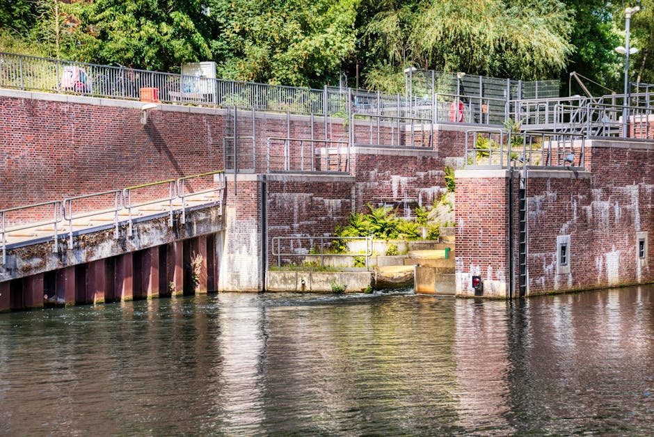 A riverside brick embankment with a metal railing and steps leading up to a walkway above, situated next to a body of water. The brick wall shows signs of weathering with some white staining and moss growth at the base. Behind the fence, trees and shrubs provide a green backdrop, while in the foreground, the water reflects the structure above. The area appears to be a designated access point for loading or unloading, possibly used during a home relocation or furniture transport process. Occasionally visible are equipment elements such as a small red light and a pipe at the water level, indicating safety or utility features. The scene is lit with natural daylight, illustrating a quiet, urban riverside environment suitable for logistical activities associated with house removals, and the presence of a corridor and stairs suggest pathways for moving furniture or packing materials during a move, supported by professional removals services like Enfield Removals.