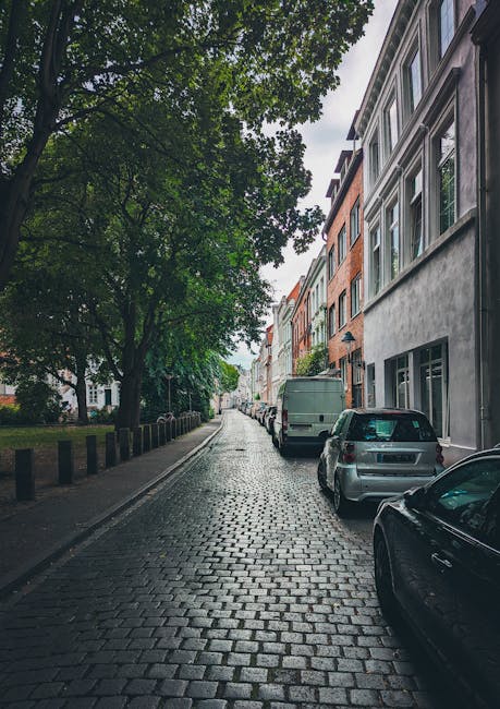 A narrow residential street in Enfield with cobblestone pavement, lined with multi-storey terraced houses featuring large windows and modern facades in neutral tones. Several parked cars, including a small grey hatchback and a larger green van, are positioned along the kerbside, indicating a typical urban setting suitable for house removals. Tall trees with lush green foliage overhang the street, providing partial shade to the parked vehicles. The overcast sky suggests diffuse lighting, creating a calm, quiet atmosphere. In the context of house removals, Enfield Removals may use such streets for loading and unloading furniture and packing materials, which are likely transported in cardboard boxes, wrapped furniture, or plastic coverings, along with equipment such as trolleys or straps, during home relocation or furniture transport activities. The scene captures a typical environment where moving services operate within narrow streets and parking restrictions.