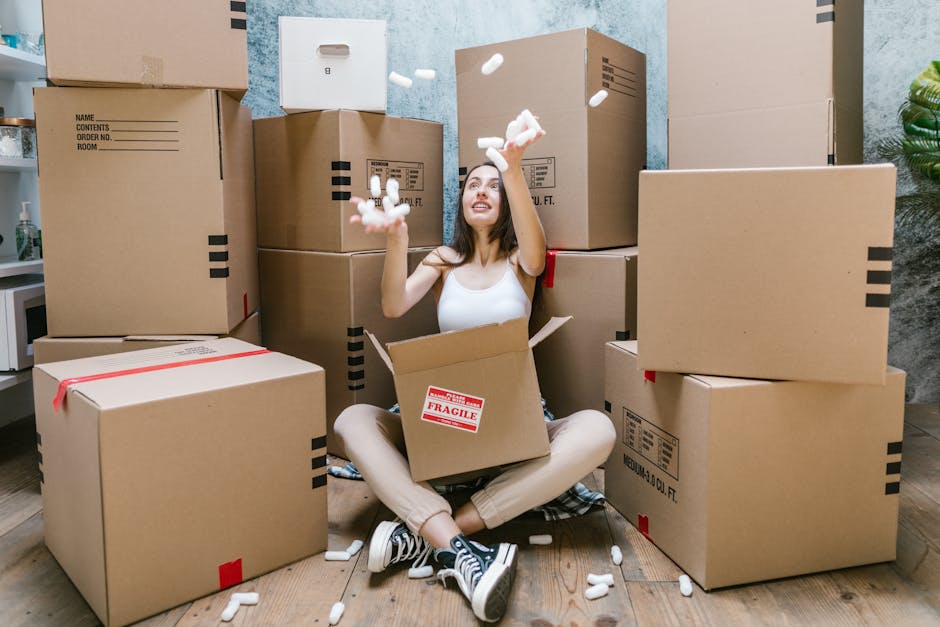 A young woman with long dark hair, sitting cross-legged on a wooden floor inside a home, surrounded by multiple large cardboard moving boxes, some sealed with red tape and others open. She is wearing a white sleeveless top, beige trousers, and black-and-white sneakers. She is smiling and looking up while throwing white packing foam peanuts into the air, with several foam peanuts scattered around her. Behind her, the wall is painted in a light blue shade, and there is a small potted plant visible on the right side of the image. The scene captures the packing and home relocation process as part of house removals, illustrating the packing stages that Enfield Removals handles during furniture transport and loading. The boxes are positioned on the floor, indicating an active packing environment typical of a professional removals service.