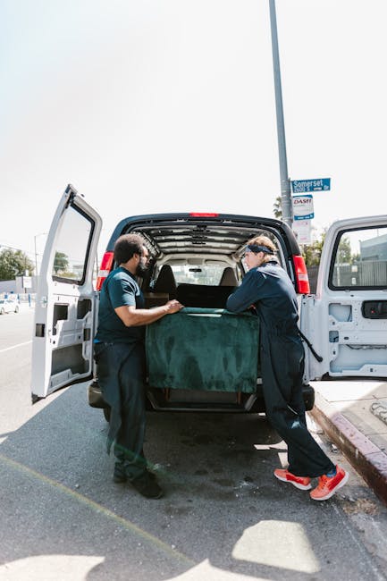 Two professional movers from Enfield Removals are engaged in the loading process at a suburban location, transferring a large, dark green upholstered furniture piece into the back of a white vans with open rear doors. The interior of the van reveals a spacious cargo area suitable for home relocation tasks, with some protective padding on the edges. One mover, dressed in dark clothing, is standing inside the van helping to stabilize the furniture, while the other, wearing a blue shirt and protective gloves, is positioned outside, guiding the item into the vehicle. Nearby, the pavement is clean and the area is well-lit by natural daylight, with a street sign and a lamppost visible in the background. This scene illustrates the careful packing and furniture transport activity involved in professional removals, highlighting the use of equipment and teamwork in the moving logistics process. Enfield Removals provides comprehensive house removals, including packing, loading, and transporting furniture for home relocations in the Enfield area.