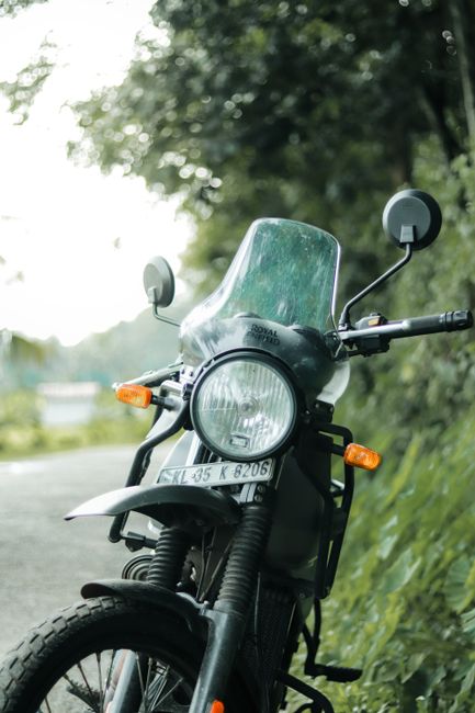 Close-up image of a black motorcycle parked on a paved roadside with a fairing windshield, rearview mirrors, and front headlight visible. The motorcycle features a UK license plate reading 'XJL 35 K 8206' and is situated next to a grassy area with green foliage and trees in the background. The scene appears to be outdoors during daylight, with natural lighting illuminating the motorcycle and surroundings. The image may be related to house removals or moving logistics, such as transport or packing procedures, and showcases the motorcycle as part of a larger move or transfer process, typical of home relocation services provided by Enfield Removals.